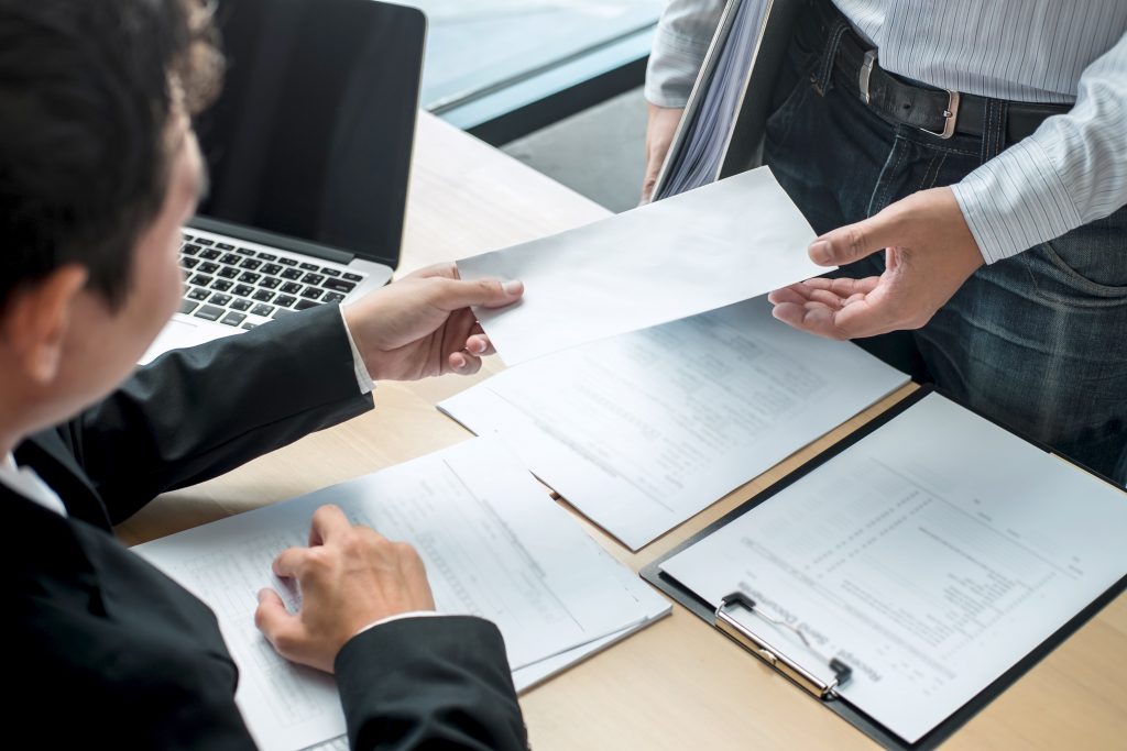 Two professionals exchange a document across a desk, with a laptop and papers nearby in a business setting.